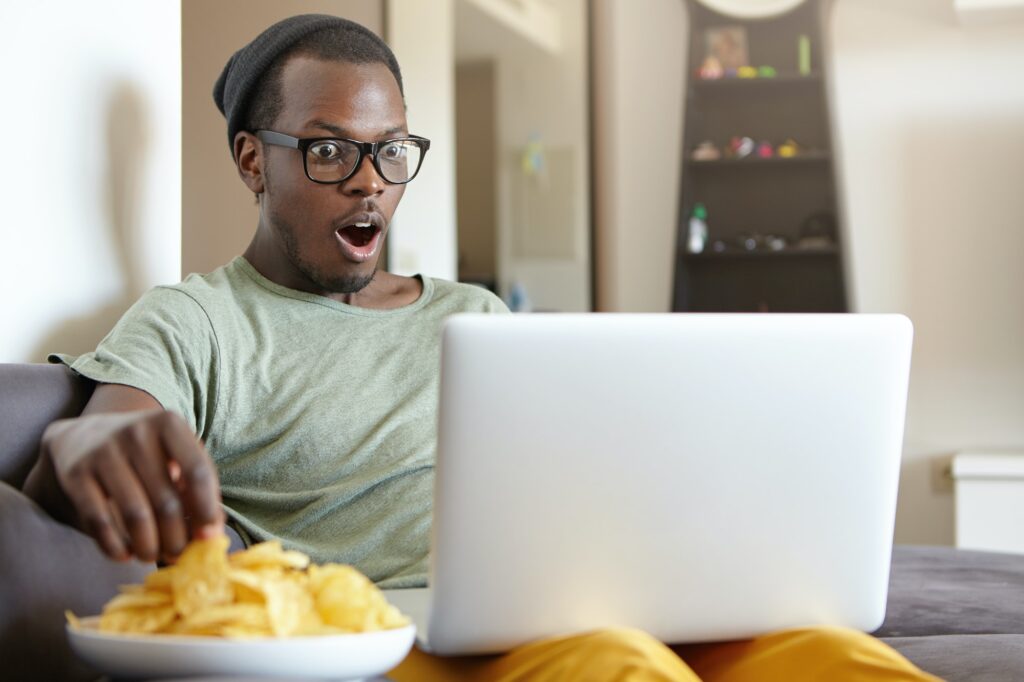 Dark-skinned guy watching movies at home over notebook eatting crisps being shocked by plot. Restful