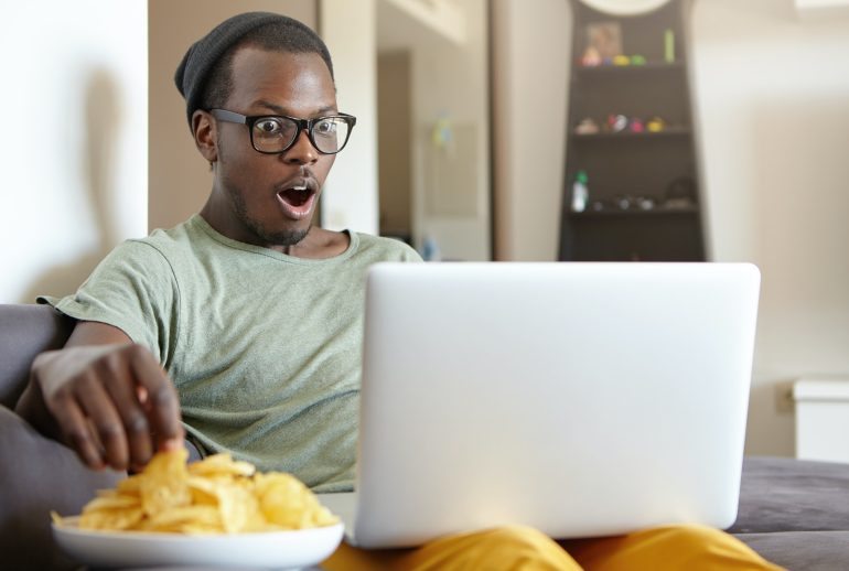 Dark-skinned guy watching movies at home over notebook eatting crisps being shocked by plot. Restful